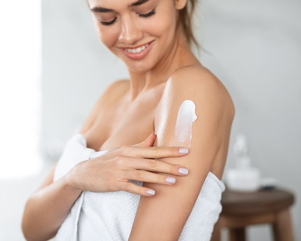 Una imagen de una chica con un toalla que envuelve el cuerpo y echándose crema en la piel del brazo. Cuidado corporal.