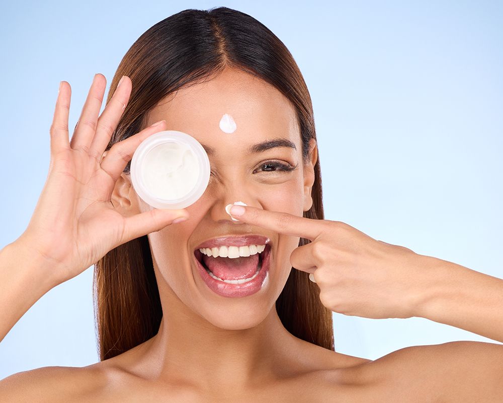 Imagen de una mujer sonriendo sujetando con una mano un bote de crema facial y con la otra aplicándose crema en la frente y en la nariz.