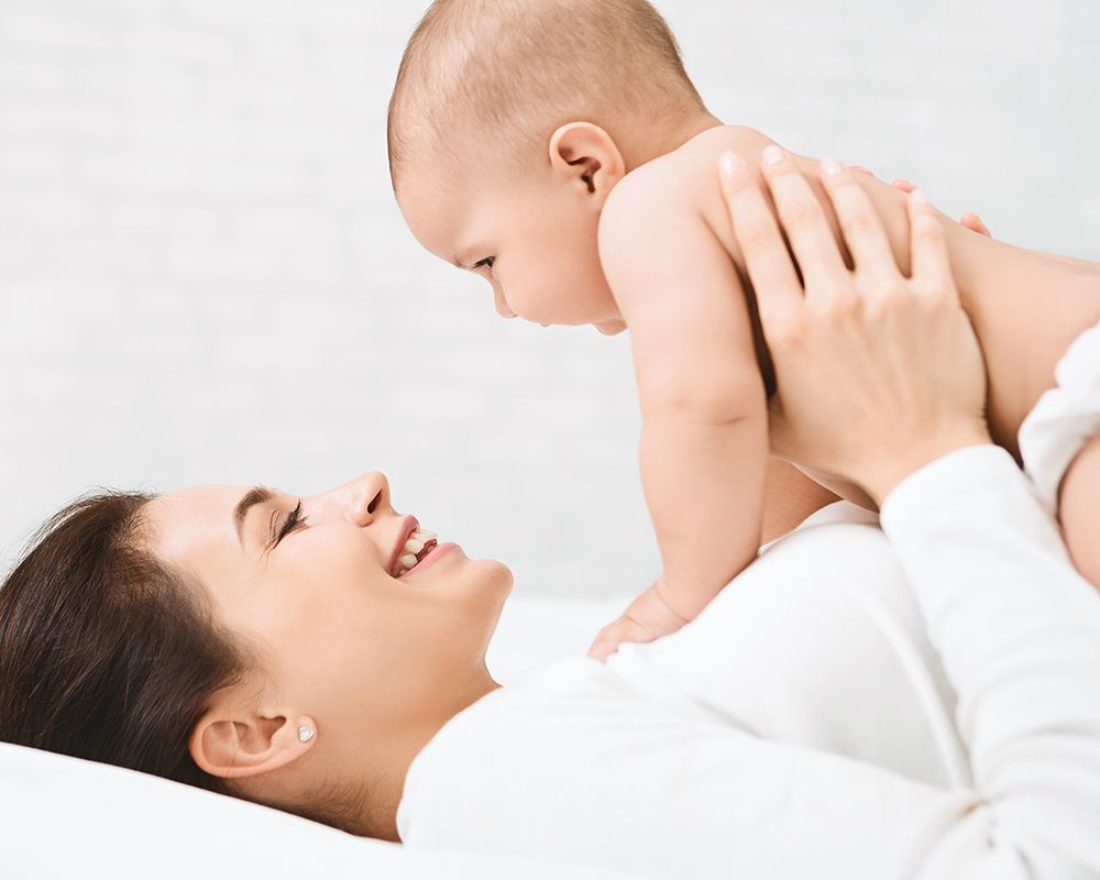 La foto muestra a un bebé sin camisa siendo sostenido por su madre. La madre está acostada, vistiendo una camiseta blanca. El fondo es de color claro, dando una sensación de tranquilidad y limpieza al ambiente. Es una imagen que hace alusión al cuidado infantil.